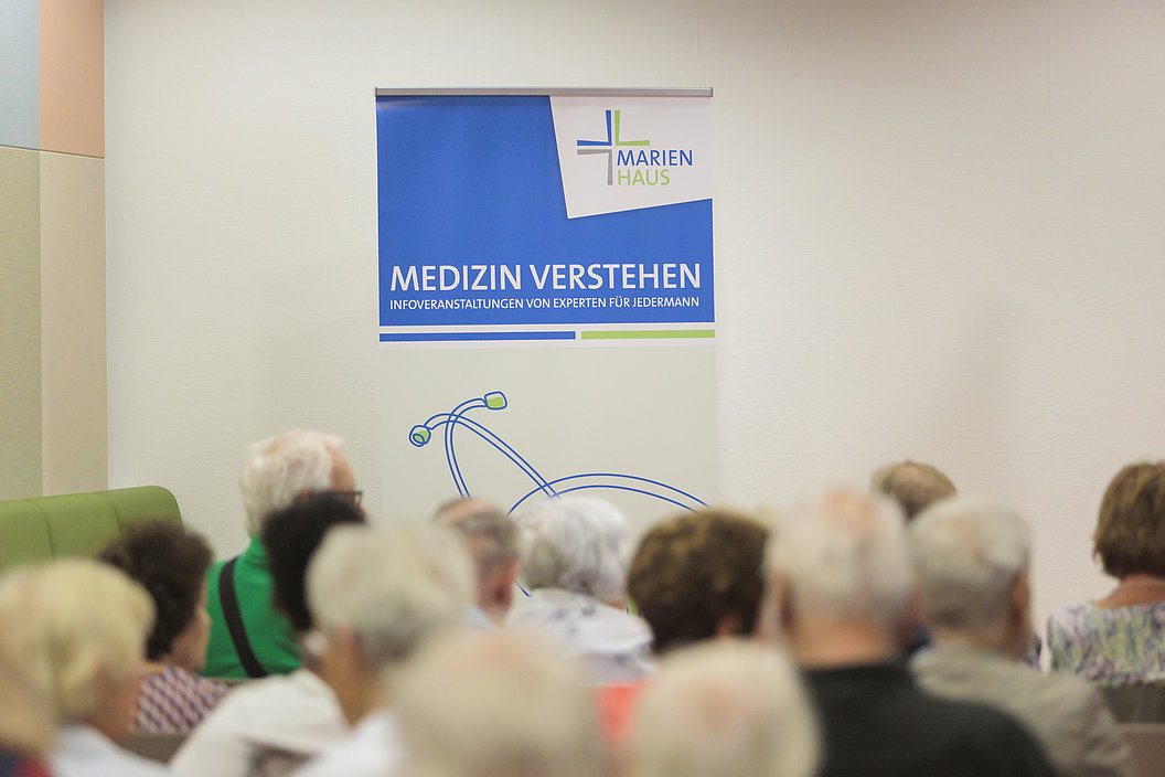 Publikum vor einem Banner mit dem Schriftzug 'MARIEN HAUS MEDIZIN VERSTEHEN INFOVERANSTALTUNGEN VON EXPERTEN FÜR JEDERMANN'