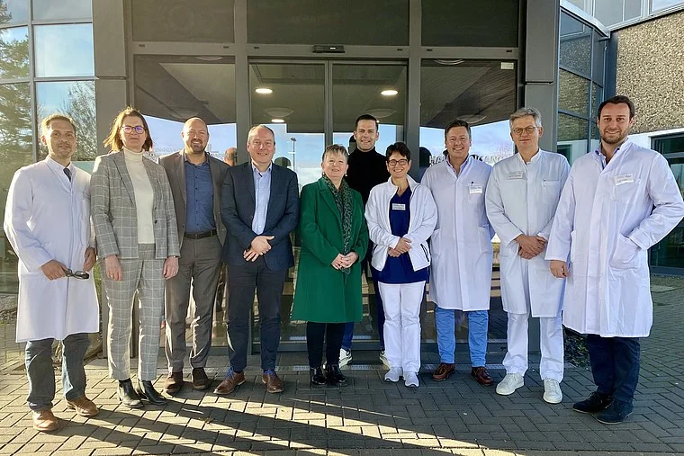 Zehn Personen stehen vor einem Gebäude, einige tragen weiße Laborkittel, andere formelle Kleidung, Gruppenfoto im Freien