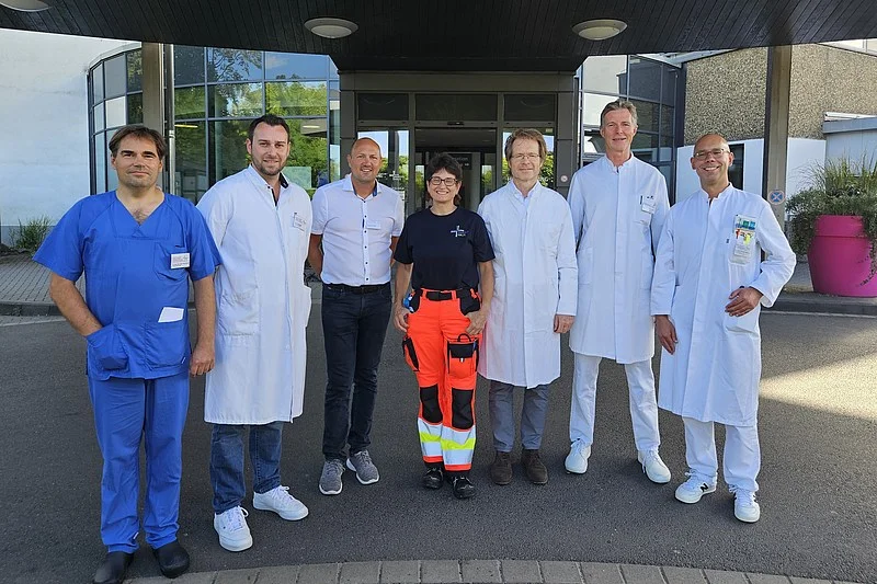 Gruppe von sieben Personen in medizinischer Kleidung, darunter Ärzte in weißen Kitteln und ein Rettungssanitäter in orangefarbener Hose, stehen vor einem Gebäude.