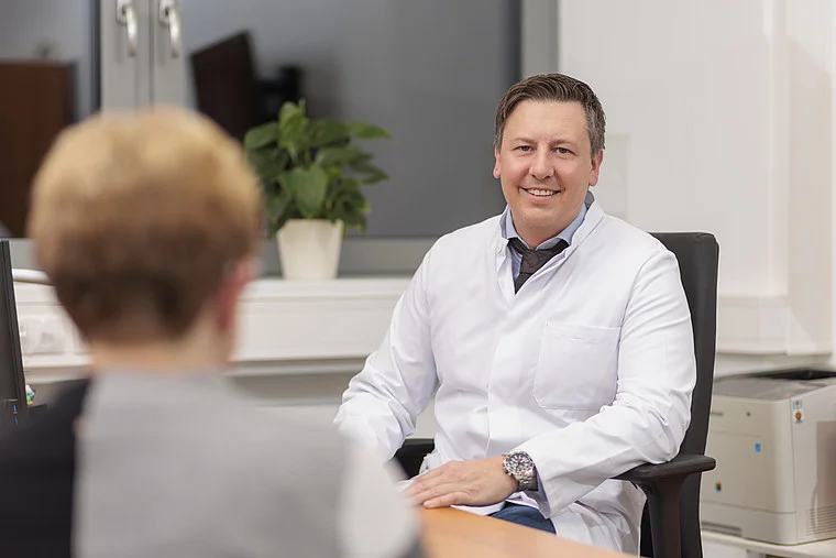 Prof. Jan Schmolders sitzt in einem weißen Laborkittel auf einem Stuhl, mit einem Tisch zwischen ihm und einer anderen Person. Im Hintergrund steht eine Pflanze.