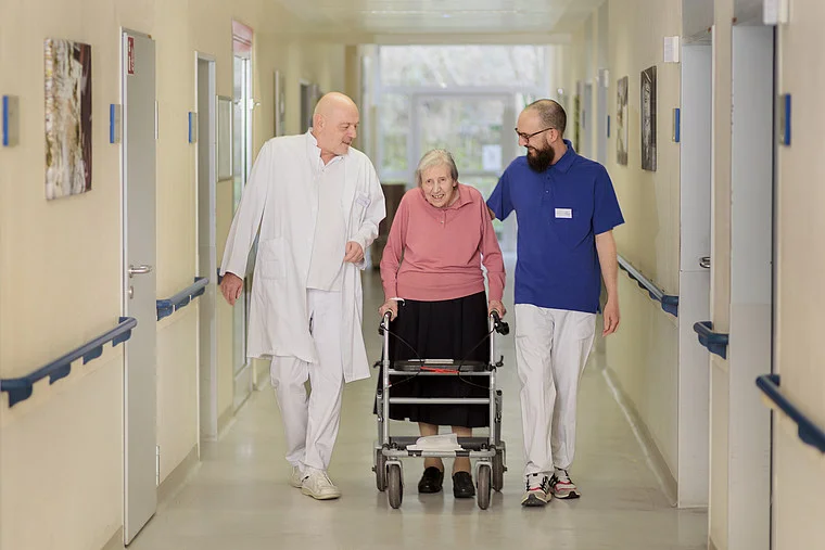 Eine ältere Frau mit einem Rollator wird von zwei Männern begleitet, einem Arzt in weißem Kittel und einem Pfleger in blauer Uniform, durch einen Krankenhausflur.