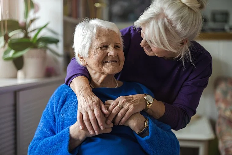Eine ältere Frau mit grauen Haaren trägt einen blauen Pullover und sitzt auf einem Stuhl. Eine andere Frau mit blonden Haaren in lila Kleidung umarmt sie von hinten.