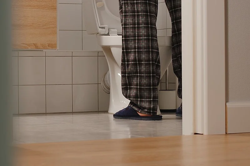 Beine einer Person in karierten Pyjamahosen und Hausschuhen vor einer geöffneten Toilettenschüssel in einem Badezimmer.
