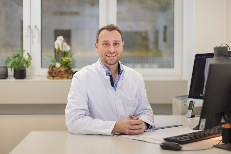 Dr. Tim Mödder sitzt in weißem Laborkittel an einem Schreibtisch mit Computer in einem hellen Büro