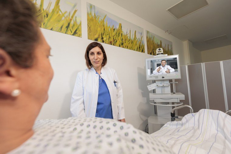 Eine Patientin liegt im Bett und schaut auf eine Ärztin in einem weißen Kittel, die vor einem Monitor steht. Der Monitor zeigt eine andere Person.